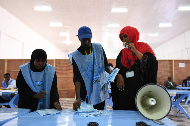 Eine Gruppe von Menschen steht um einen Tisch mit einem Megafon, Papieren und anderen Gegenständen, mit einigen Menschen im Hintergrund sitzend und stehend, unter einer Decke mit Lampen, an einer Wahllokal.