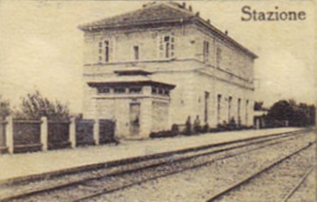 Altes Schwarz-Weiß-Foto eines Bahnhofsgebäudes in der Mitte von Bäumen umgeben, mit einem Zaun auf der linken Seite, Eisenbahnschienen unten und Text oben.