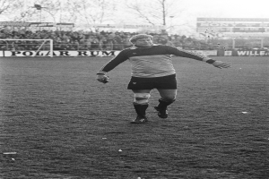 Ein Mann in einem Fußballtrikot steht auf einem Feld, umgeben von einer Menge, Torpfosten, Planken und Bäumen unter einem klaren Himmel.