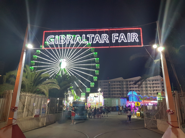 Eintritt zum Gibraltarer Volksfest bei Nacht mit beleuchtetem Riesenrad und Namensschild, Menschenmenge, Bäumen und Gebäuden im Hintergrund vor dunklem Himmel.