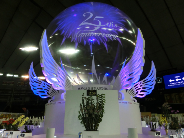 Ein großer Kristallball mit Flügeln oben drauf, umgeben von Blumenbouquets, Namensschildern und anderen Gegenständen auf einem dekorierten Boden, mit Schildern, Lichtern und einem Bildschirm im Hintergrund.