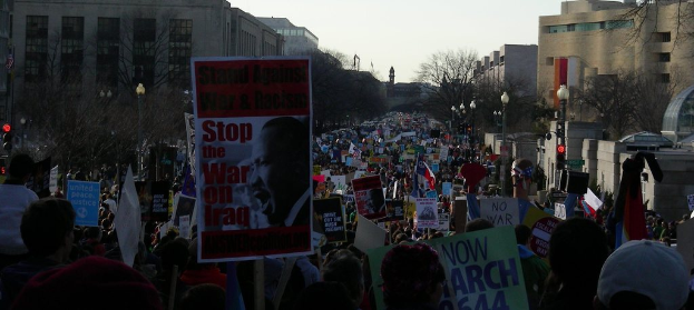 Eine Gruppe von Menschen demonstriert auf einer Straße, hält Plakate, mit Gebäuden, Bäumen, Laternen und einem klaren Himmel im Hintergrund.