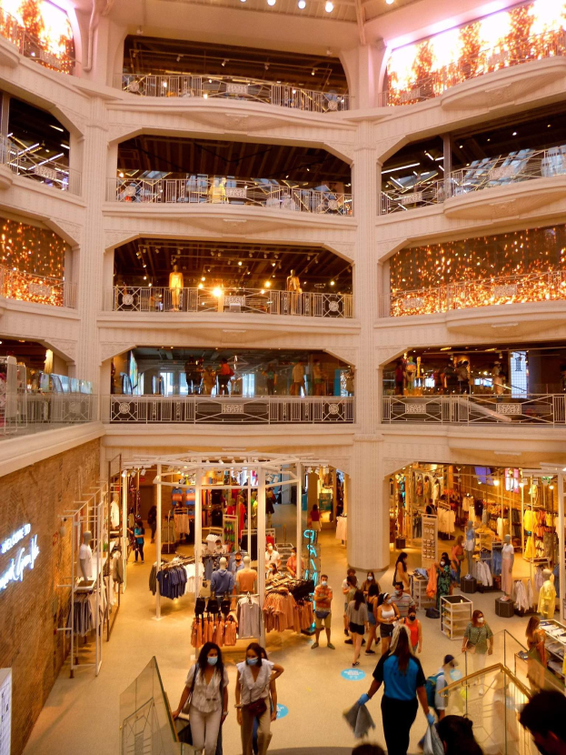 Großer Atrium eines belebten Einkaufszentrums mit Menschen, die Taschen tragen und Schaufensterpuppen mit Kleidern anschauen, umgeben von Geländern, Beleuchtung und Texttafeln an den Wänden.