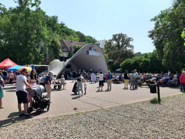 Eine Menschenmenge steht vor einem Gebäude bei einer Fest-Eröffnung, einige sitzen auf Bänken, andere stehen; ein Kinderwagen ist im Vordergrund, und Bäume, Gebäude und ein klarer blauer Himmel sind im Hintergrund zu sehen, mit Zelten links und einem Müllcontainer rechts.