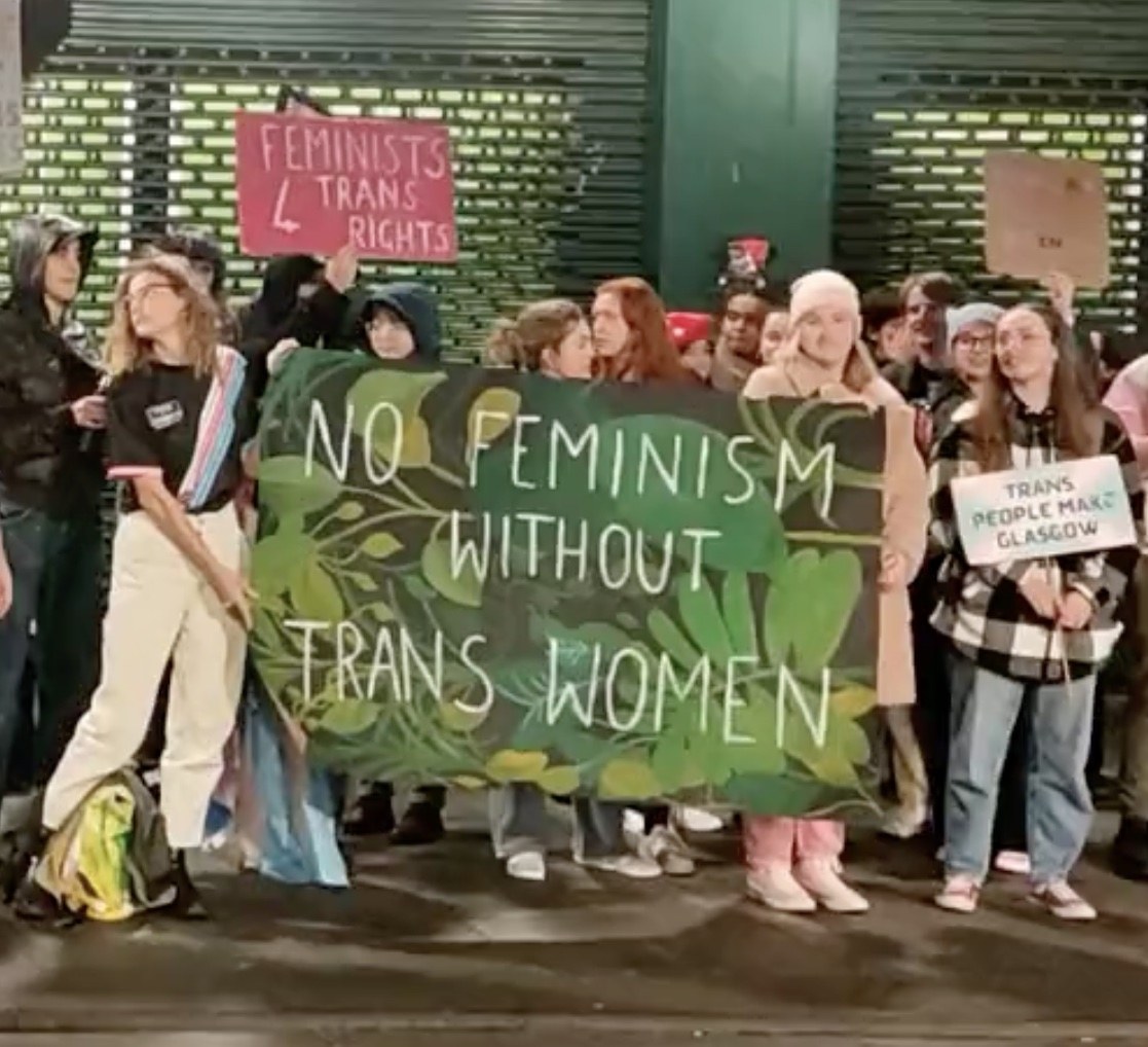 Eine Gruppe von Menschen hält ein Banner mit der Aufschrift "Kein Feminismus ohne Trans-Frauen" und verschiedene Plakate in einem öffentlichen Raum.
