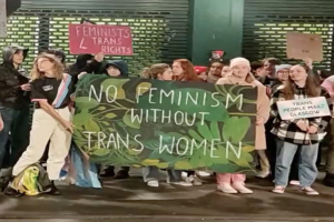 Eine Gruppe von Menschen hält ein Banner mit der Aufschrift "Kein Feminismus ohne Trans-Frauen" und verschiedene Plakate in einem öffentlichen Raum.
