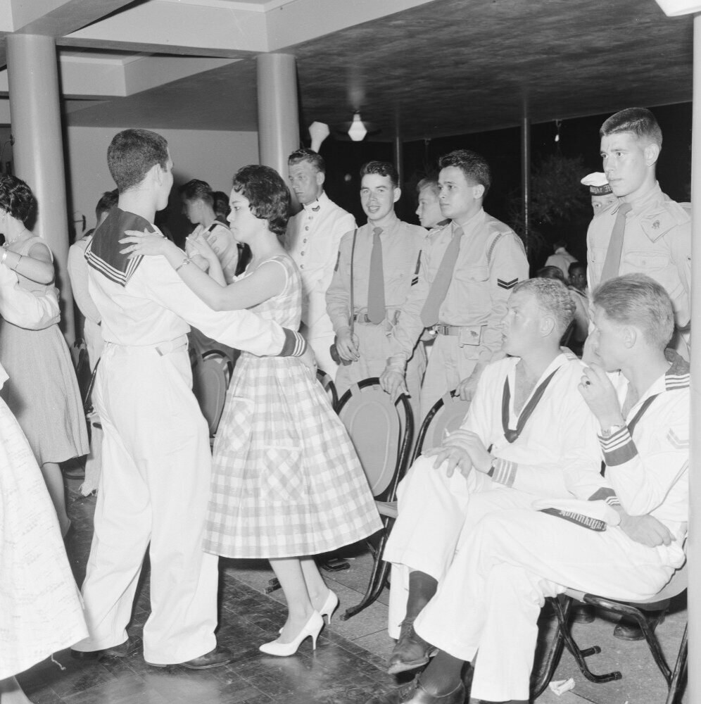 Schwarz-weiß-Bild einer Gruppe von Männern und Frauen in Militäruniformen, die auf einem Tanzboden tanzen und auf Stühlen sitzen, mit Säulen und Lichtern im Hintergrund.