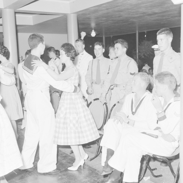 Schwarz-weiß-Bild einer Gruppe von Männern und Frauen in Militäruniformen, die auf einem Tanzboden tanzen und auf Stühlen sitzen, mit Säulen und Lichtern im Hintergrund.