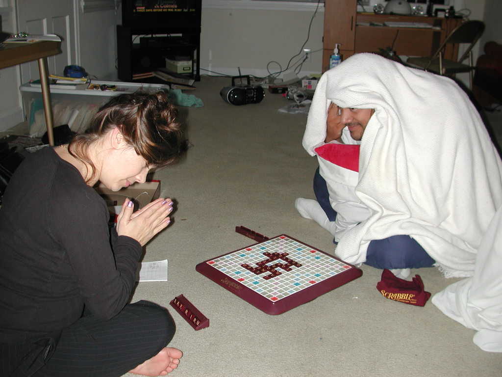 Zwei Personen sitzen auf dem Boden und spielen ein Spiel; die Person links trägt ein schwarzes Kleid, die Person rechts trägt Weiß, mit einem Stuhl und einem Tisch dahinter und Kabel auf dem Boden.