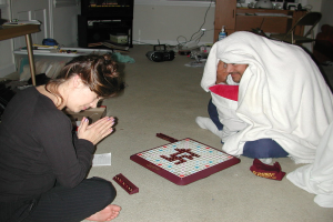 Zwei Personen sitzen auf dem Boden und spielen ein Spiel; die Person links trägt ein schwarzes Kleid, die Person rechts trägt Weiß, mit einem Stuhl und einem Tisch dahinter und Kabel auf dem Boden.