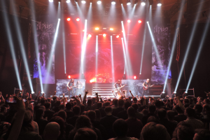 Eine Gruppe von Menschen, die auf einer hell erleuchteten Bühne spielen, während andere das Konzert mit ihren Handys aufnehmen.