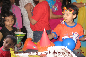 Kinder vor einem Tisch mit Kuchen, Luftballons und Mützen, hinter ihnen Erwachsene