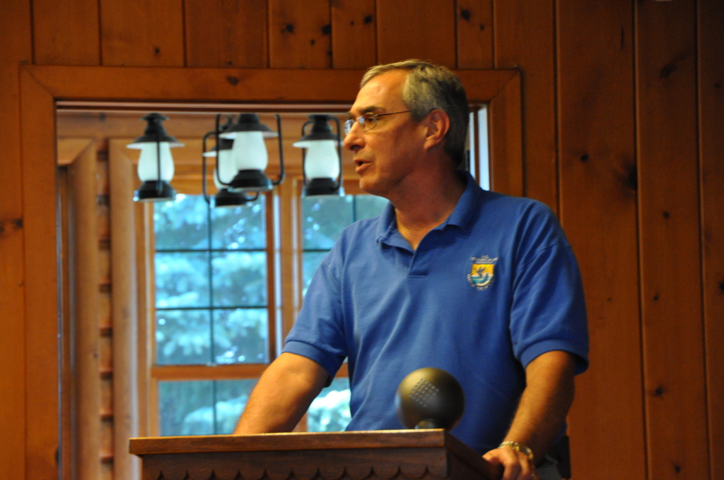 Eine Person in einem blauen T-Shirt steht hinter einem hölzernen Rednerpult in einem Raum, mit einer Wand, einem Fenster und hängenden Lampen im Hintergrund.
