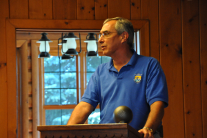 Eine Person in einem blauen T-Shirt steht hinter einem hölzernen Rednerpult in einem Raum, mit einer Wand, einem Fenster und hängenden Lampen im Hintergrund.