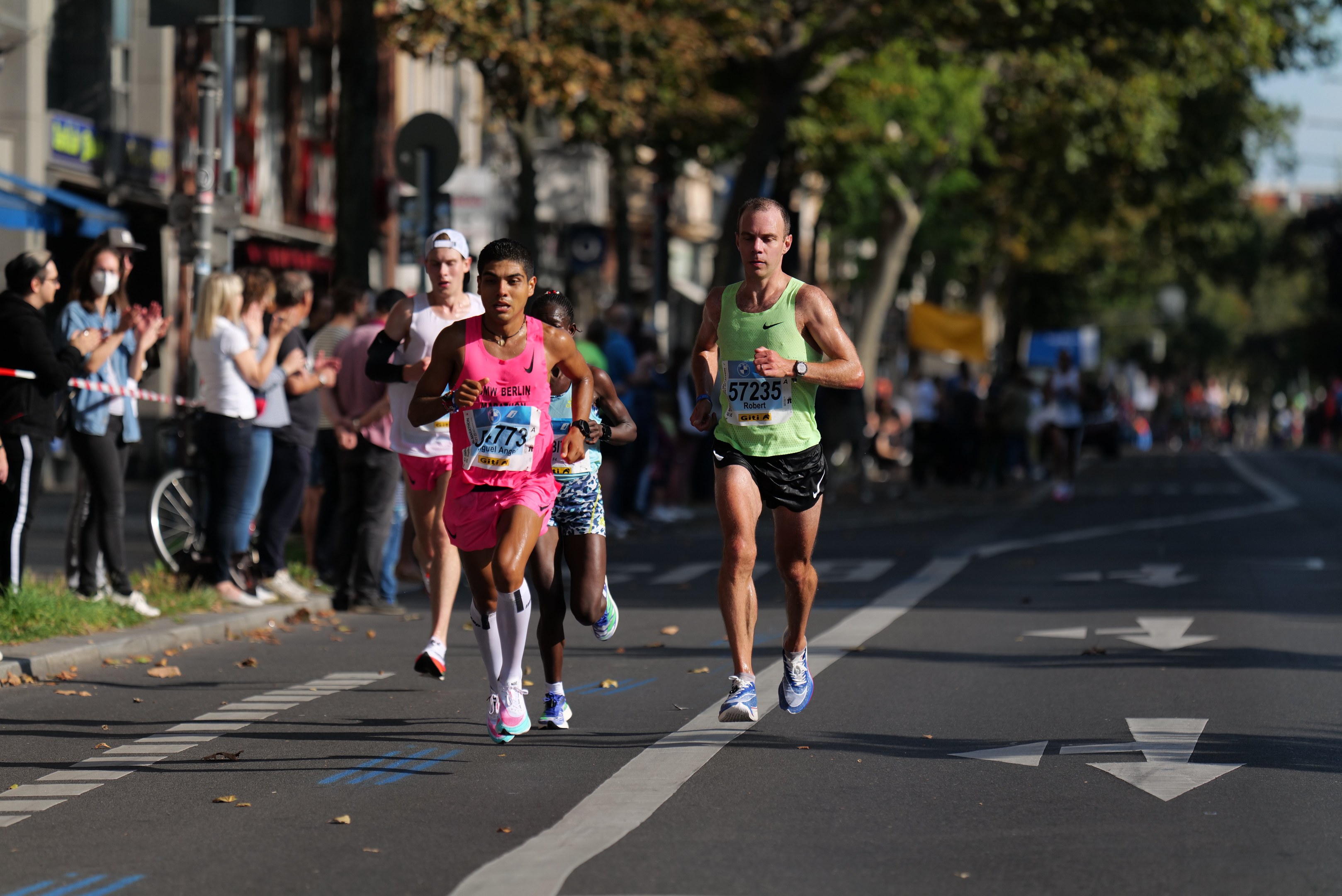 Gruppe von Läufern bei einem Stadtmarathon, mit Zuschauern auf der linken Seite, unscharfen städtischen Hintergrund mit Bäumen, Gebäuden und einem Fahrrad.