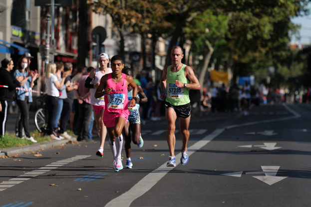Gruppe von Läufern bei einem Stadtmarathon, mit Zuschauern auf der linken Seite, unscharfen städtischen Hintergrund mit Bäumen, Gebäuden und einem Fahrrad.