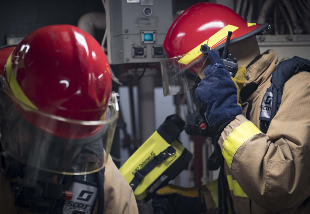 Zwei Feuerwehrleute in Schutzausrüstung bei der Arbeit an einem Feuerhydranten während einer übung mit Gerät und Kabeln im Hintergrund.