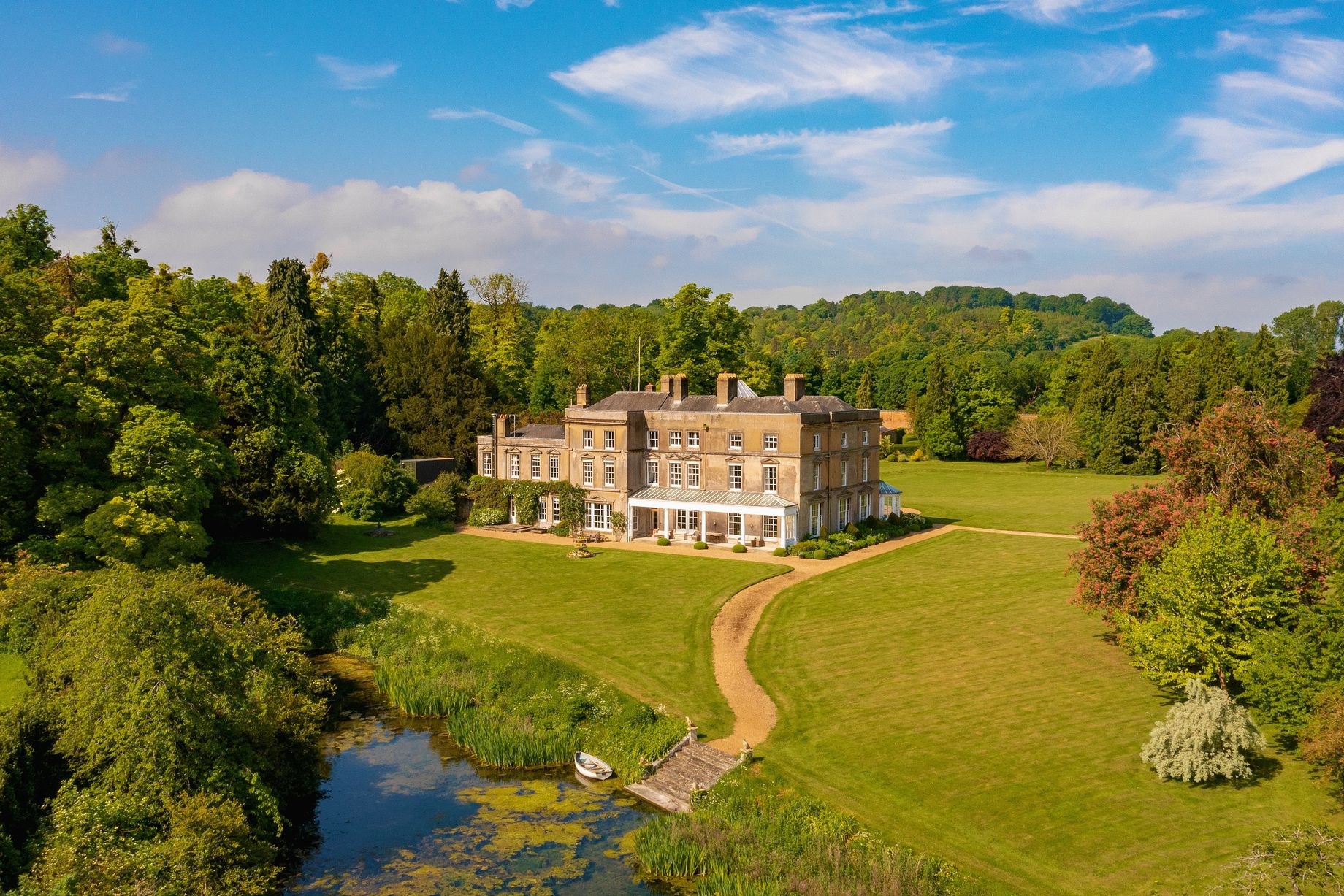 Luftaufnahme eines großen, teuren Anwesens in einer grünen Landschaft mit einem Weg, umgeben von Bäumen und Pflanzen, einem Gewässer im Vordergrund und bewölktem Himmel.