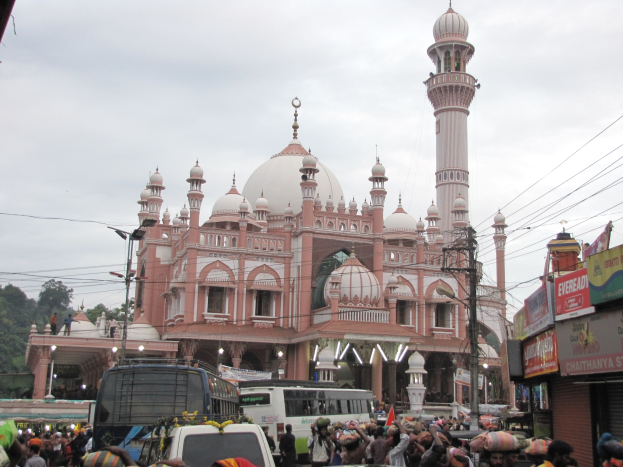 Eine große Menschenmenge geht eine Straße vor einer Moschee entlang, mit Fahrzeugen, Schirmen, Gebäuden mit Schildern, Strommasten mit Drähten, Bäumen und einem klaren blauen Himmel im Hintergrund.