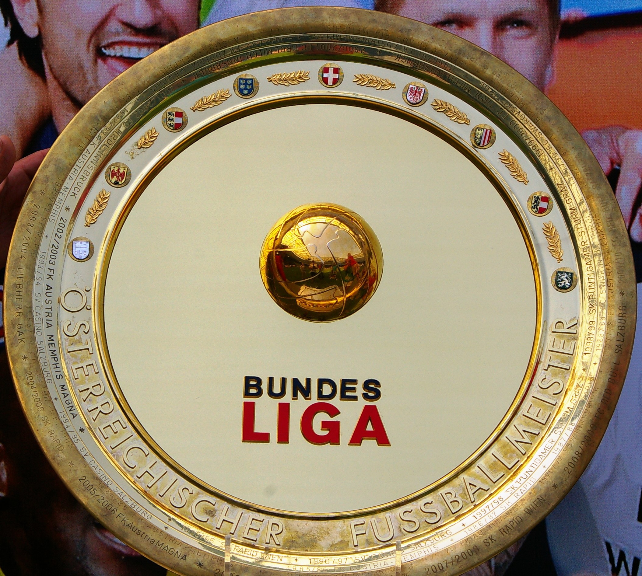 Ein Mann, der einen goldenen Schild mit der Aufschrift "Bundesliga" hält, mit einem Banner im Hintergrund, auf dem drei Personen zu sehen sind.