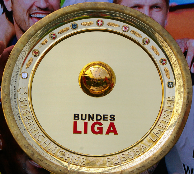 Ein Mann, der einen goldenen Schild mit der Aufschrift "Bundesliga" hält, mit einem Banner im Hintergrund, auf dem drei Personen zu sehen sind.