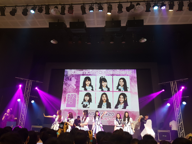 Eine Gruppe von Menschen, die auf einer hell erleuchteten Bühne spielen, mit einer Menge darunter, mit einem Sänger mit Mikrofon und einem Musiker, der ein Instrument spielt, und einem großen Bildschirm im Hintergrund, der Bilder von BK48 zeigt.