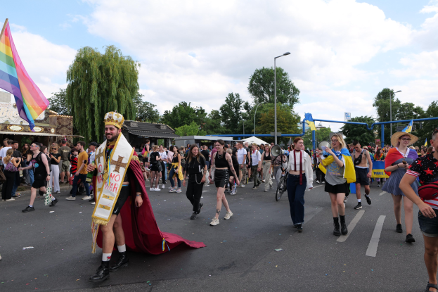 Eine Gruppe von Menschen marschiert auf der 2018er Gay Pride Parade mit einer Regenbogenfahne und Musikinstrumenten, im Hintergrund sind Laternenpfähle, Bäume und Hütten zu sehen.