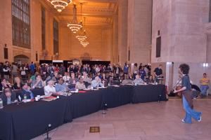 Eine Konzertszenen mit einer Menschenmenge, einem Mann, der auf der rechten Seite Gitarre spielt und singt, einer Wand und Fenstern im Hintergrund, Lichtern oben und einem Boden unten.