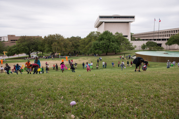 Gruppe von Menschen beim Ostereier-Spiel auf einer grünen Wiese mit Bäumen, Gebäuden, Fahnen, einem Brunnen und einem bewölkten Himmel.