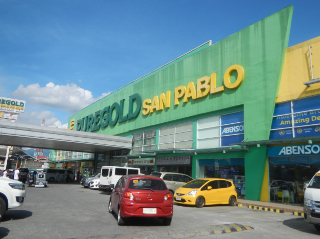 Eine belebte Stadtstraße mit Gebäuden, Fahrzeugen, Fußgängern, Straßenmöblierung, einer Brücke und einem vor einem San Pablo Geschäft geparkten Auto.