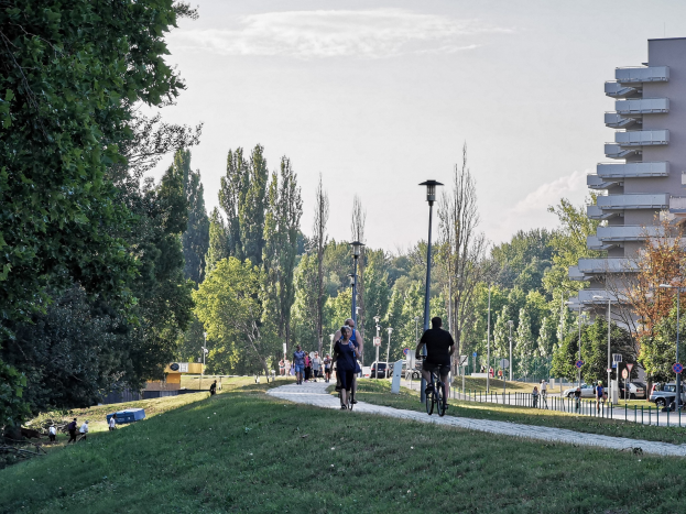 Eine Gruppe von Menschen, die auf Fahrrädern eine Parkweg mit Gras, Bäumen, Straßeninfrastruktur, Fahrzeugen und Gebäuden im Hintergrund unter einem bewölktem Himmel fahren.