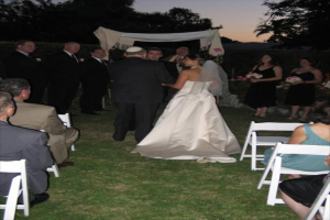Eine Gruppe von Menschen steht bei einer Hochzeit, wobei die Frauen auf der rechten Seite Blumensträuße halten, während andere auf Stühlen sitzen, umgeben von Bäumen.