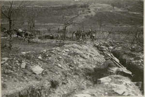 Eine Gruppe von Soldaten in Helmen und mit Gewehren marschiert auf einer Schotterstraße entlang, umgeben von Bäumen und Hügeln im Hintergrund; Text ist unten im Bild zu sehen.