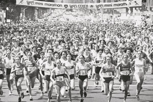 Eine Gruppe von Läufern bei einem Marathon, mit Zuschauern an der Straße, unter einem Banner mit Text, in einem Schwarz-Weiß-Bild.