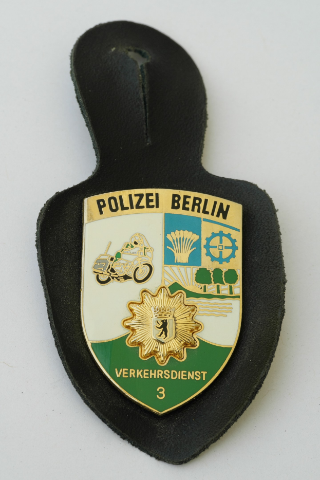 Ein schildförmiges Polizeibadged mit "Polizei Berlin" in schwarzer Schrift, mit einem blauen Hintergrund, weißem Rand und einem zentralen weißen Stern, das auf einer Oberfläche liegt.