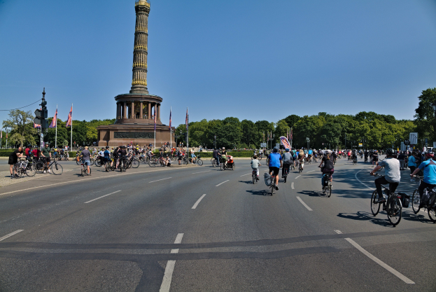 Eine große Gruppe von Menschen auf Fahrrädern, die eine Straße entlangfahren, mit einem Denkmal und Fahnen im Hintergrund, Bäumen, die die Straße säumen, und dem Himmel darüber