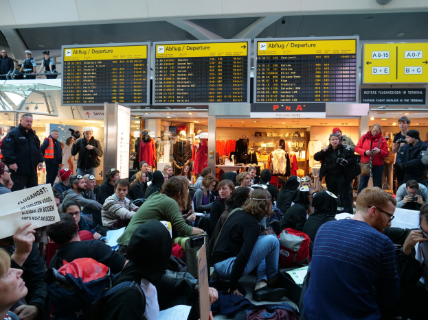 Eine große Gruppe von Menschen an einem Flughafen, einige sitzen mit Taschen und Papieren, andere stehen, mit Texttafeln, Schaufensterpuppen in Kleidern und Deckenbeleuchtung im Hintergrund, was auf eine Demonstration hinweist.