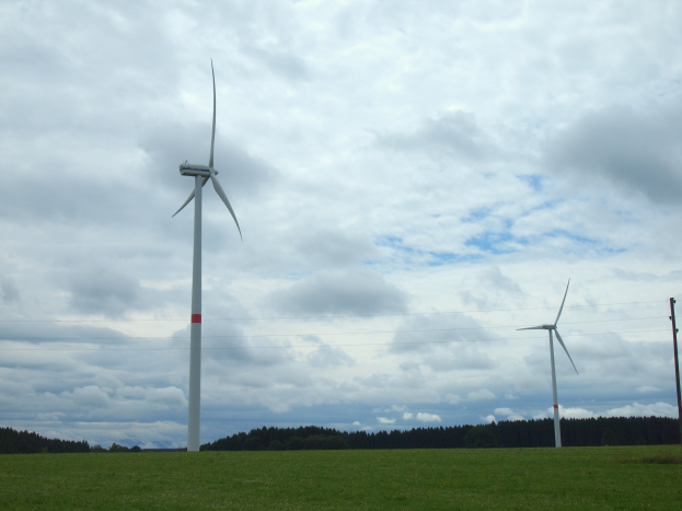 Drei weiße Windräder stehen hoch in einem grünen Feld mit Bäumen im Hintergrund und Wolken am Himmel.