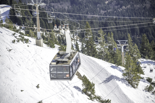 Eine mit Menschen gefüllte Seilbahn fährt einen schneebedeckten Berg hinauf, mit hohen, grünen Bäumen im Hintergrund.