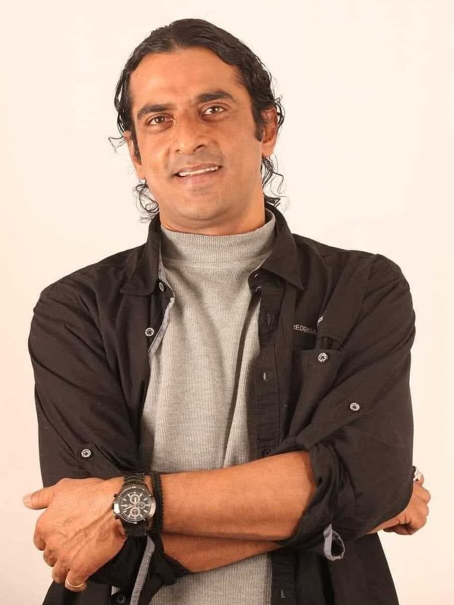 Ein Mann in einem schwarzen und aschfarbenen Kleid mit überkreuzten Armen, der eine Uhr trägt und gegen einen weißen Hintergrund steht, der tief nachdenklich erscheint.