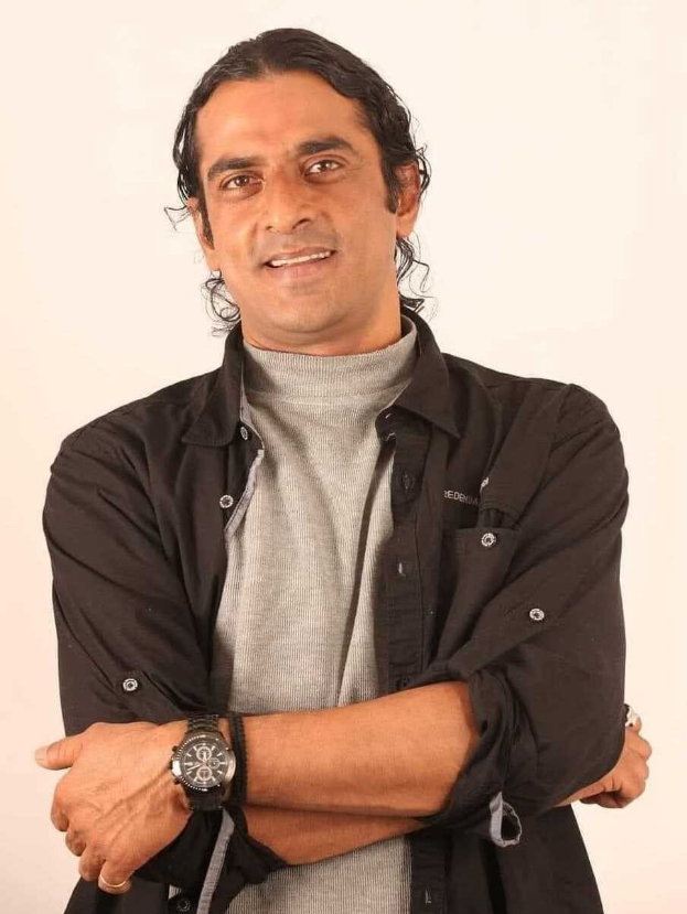 Ein Mann in einem schwarzen und aschfarbenen Kleid mit überkreuzten Armen, der eine Uhr trägt und gegen einen weißen Hintergrund steht, der tief nachdenklich erscheint.