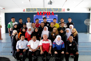 Eine Menschenmenge, die fröhlich posiert und vor einer Bühne fotografiert wird, mit einem Schild an der Wand im Hintergrund, einem Tischventilator rechts, einem Podium links und einem Projektor oben auf dem Bild.