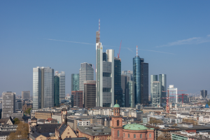 Die Frankfurter Skyline von einem Hügel aus betrachtet, mit einem prominenten Wolkenkratzer in der Mitte umgeben von verschiedenen Gebäuden und Bäumen, mit dem Himmel im Hintergrund.