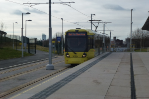Eine Straßenbahn auf ihren Schienen mit Straßenlaternen, Strommasten, einem Eisengitter und einer Plattform im Vordergrund, mit Gebäuden, Bäumen und dem Himmel im Hintergrund.