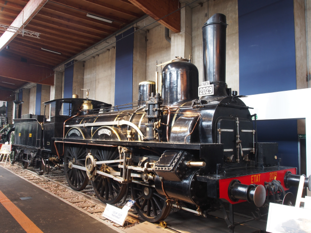 Eine gro├če, alte Dampflokomotive in einem Museums Setting umgeben von Eisenbahnschienen, Informationsschildern und anderen Ausstellungen, mit einer beleuchteten Decke und Wand im Hintergrund.