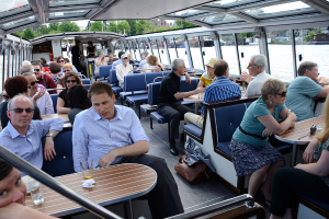 Ein Bootinneres mit Sitzreihen, die von Menschen besetzt sind, Tischen mit Gläsern und Schüsseln davor, einer Tasche auf dem Boden, Wasser im Hintergrund, Gebäuden auf der rechten Seite und kleinen Pflanzen über dem Boot.