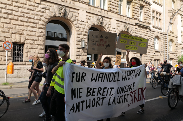 Eine Gruppe von Menschen, die auf einer Stadtstraße marschieren, einige halten Schilder und andere fahren Fahrräder, im Hintergrund ein Gebäude mit Rundbogenfenstern, Säulen und Skulpturen.