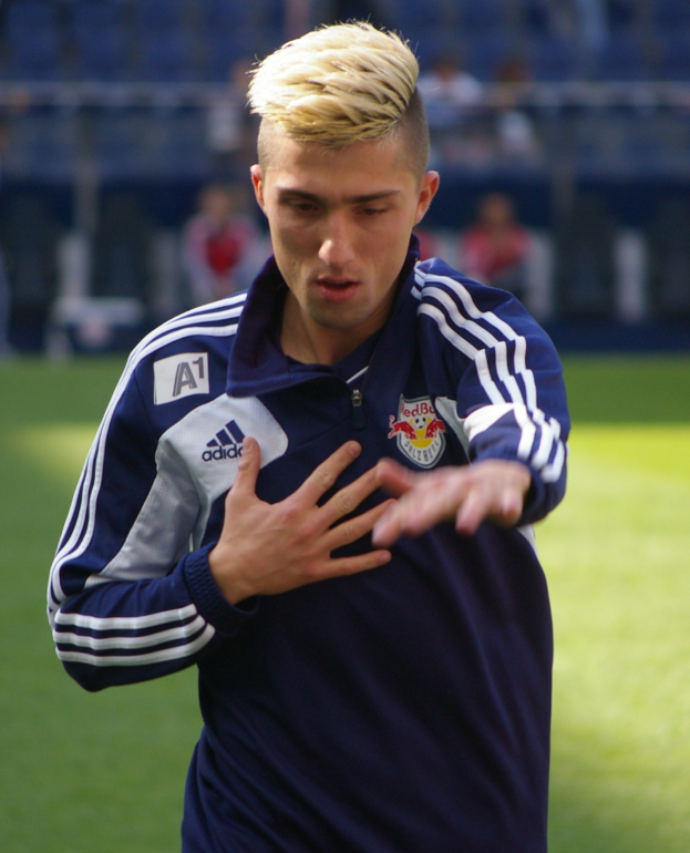 Ein junger Mann in einem blauen T-Shirt steht auf einem Fußballfeld, die Arme ausgestreckt, mit entschlossener Miene, während der Hintergrund unscharf ist.