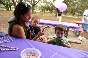 Eine Frau in Schutzbrille hält ein Baby, mit einem Jungen in einem grünen Kleid in der Nähe, der neben Tischen mit einer Geigenpartitur und einer Schale mit Würfeln, Ballons und einem Mann auf der rechten Seite steht, mit Bäumen im Hintergrund.