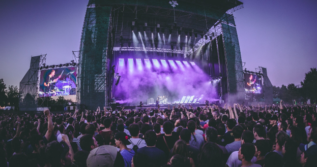 Eine große Menschenmenge bei einem Musikfestival schaut auf der Bühne mit Instrumenten und Bildschirmen, die von einem Gebäude, Bäumen und einem klaren blauen Himmel umgeben sind.